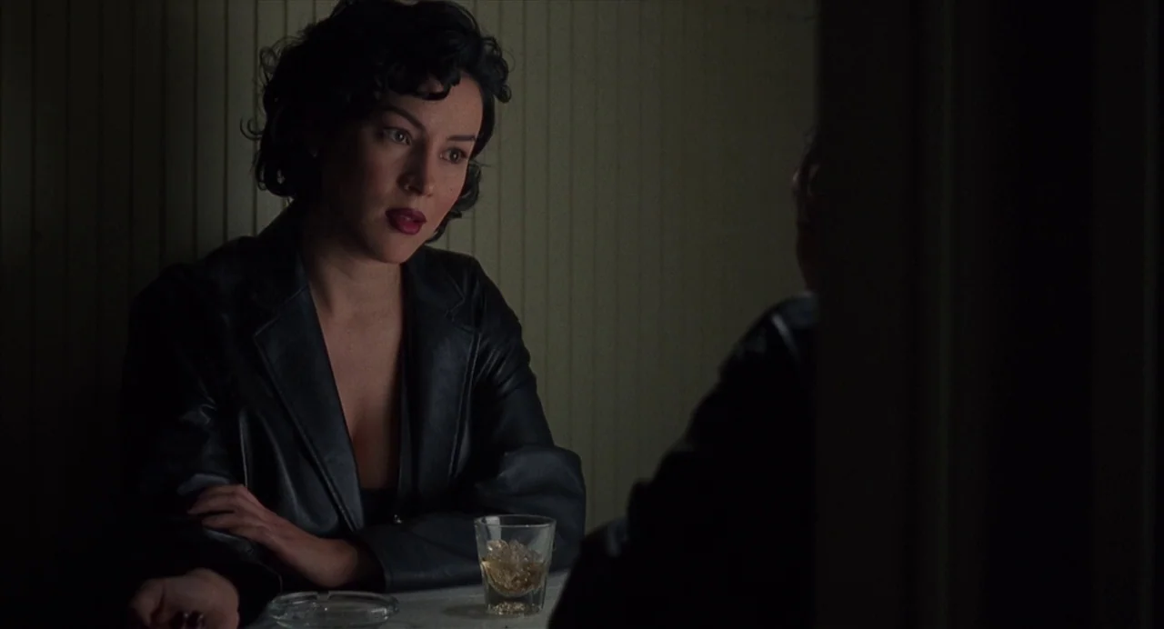 Bound (1996) movie still: medium — A dimly lit restaurant booth with vertical wood slat wall paneling. A woman in a black leather jacket sits across…