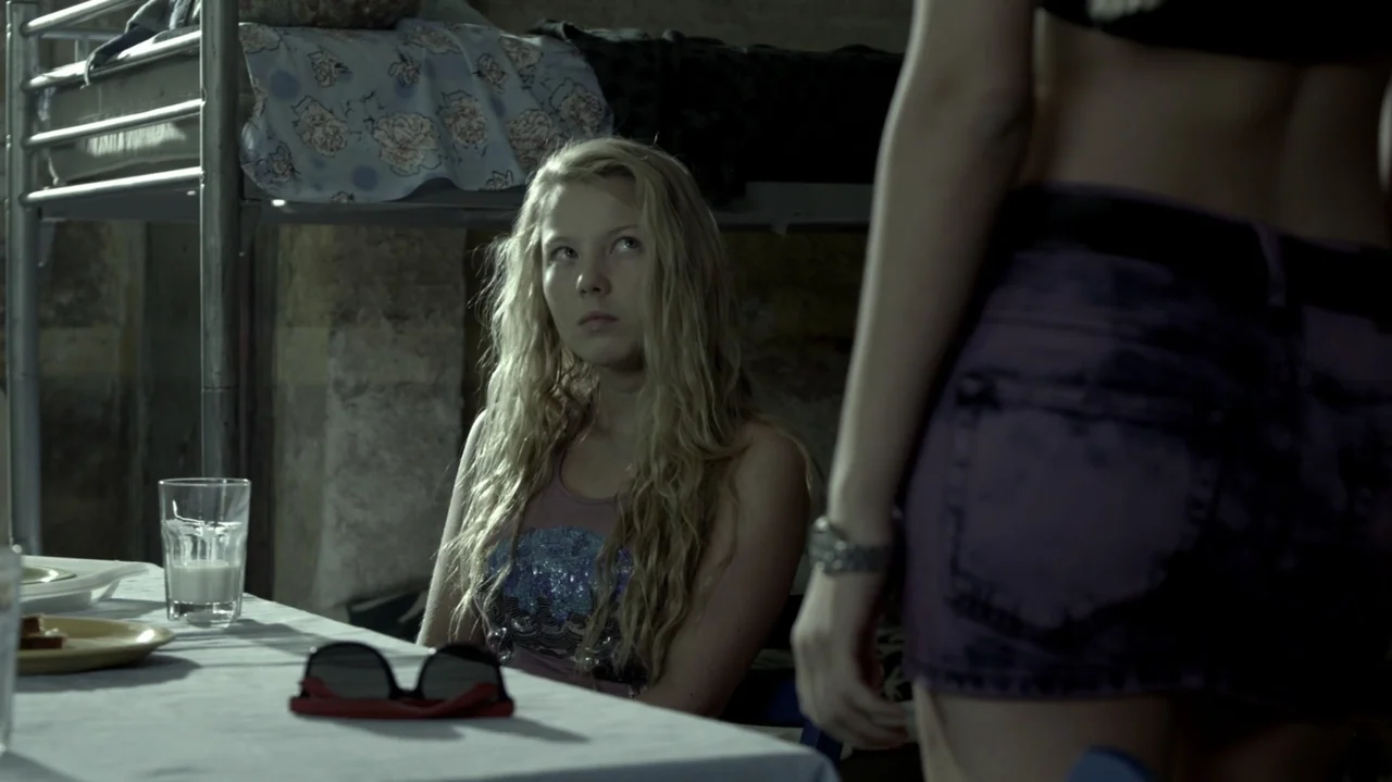 6 Bullets (2012) movie still: medium — A dimly lit room contains a metal bunk bed and a table with glassware. A young blonde woman sits at the table…