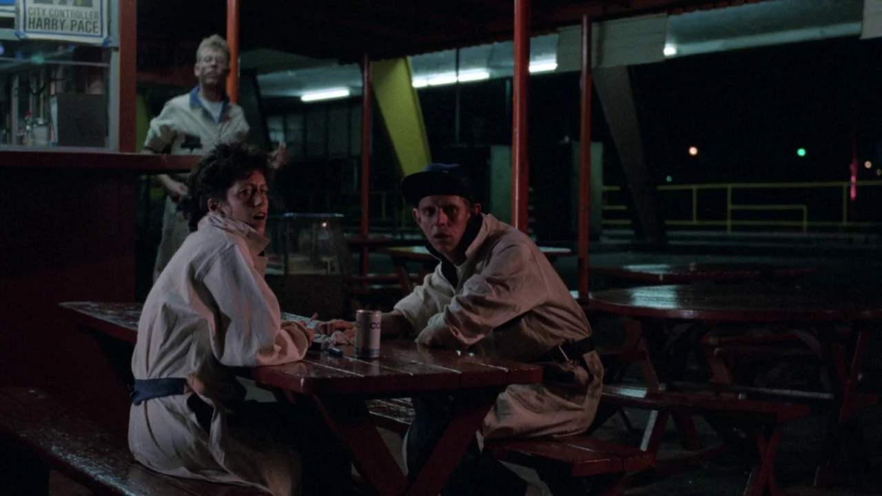 Repo Man (1984) movie still: medium — A nighttime outdoor picnic area features red wooden tables under long fluorescent lights. Two individuals sit…