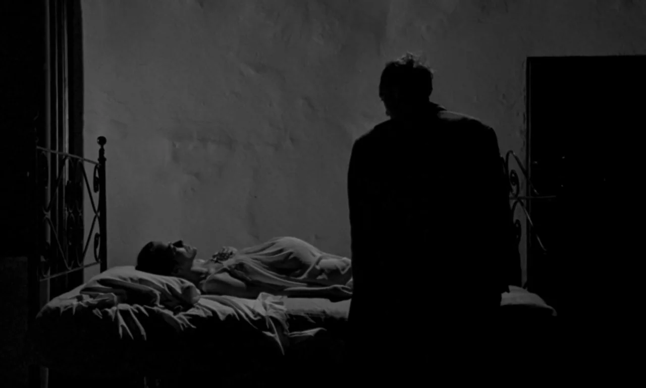 Nostalghia (1983) movie still: medium — A dark bedroom with textured walls features a metal-frame bed against one side. A man stands silhouetted…