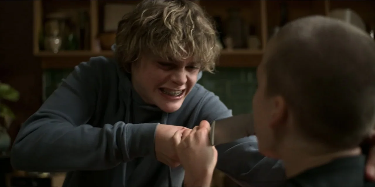 Bring Her Back (2025) movie still: medium — A dimly lit room with wooden cabinets visible in the soft-focused background. A young man with braces…