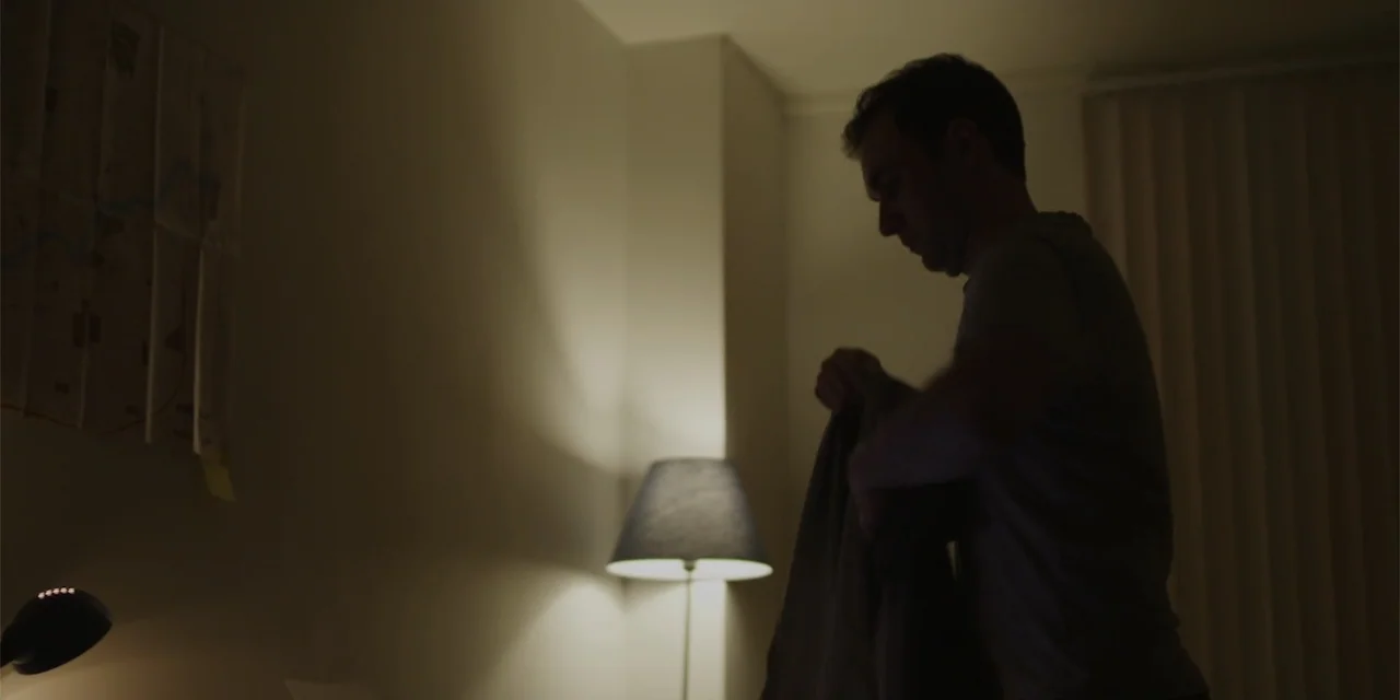Lobos (2019) movie still: medium — A dimly lit bedroom features a floor lamp and a wall map in shadow. A man is silhouetted as he prepares to put on…