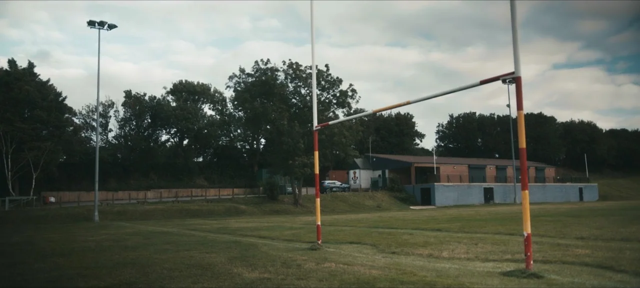 No Woman No Try (2022) movie still: wide — An empty outdoor rugby pitch features a large H-shaped goalpost in the foreground. A long brick and…