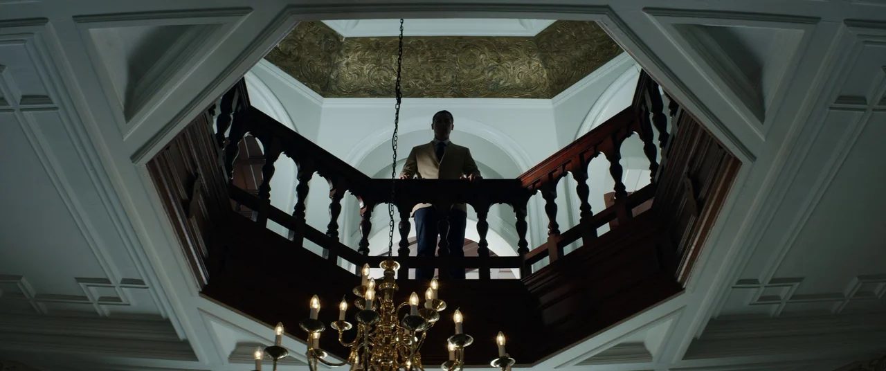 The Empty Man (2020) movie still: wide — An opulent stairwell with carved wooden railings and a gold-painted octagonal ceiling. A man in a tan…