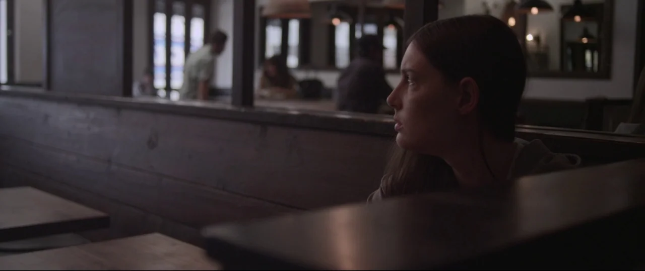 Sun Choke (2015) movie still: medium — A dimly lit restaurant interior features dark wooden booths with blurred patrons in the distance. A young…