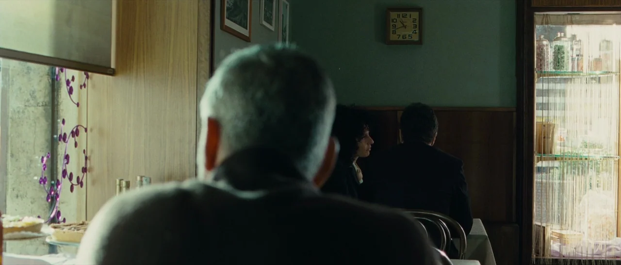 The American (2010) movie still: medium — A dimly lit diner with wooden paneling and a large window facing the street. Two people sit at a distant…