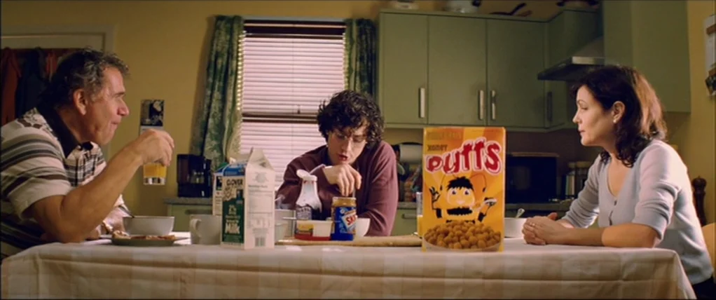 Kick Ass (2010) movie still: medium — A residential dining area features a table with a plaid tablecloth and cereal boxes. Three family members sit…
