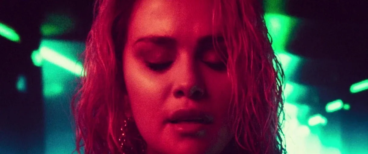 Emilia Pérez (2024) movie still: closeup — A nightclub environment filled with blurred green neon lights. A young woman with damp hair stands with…