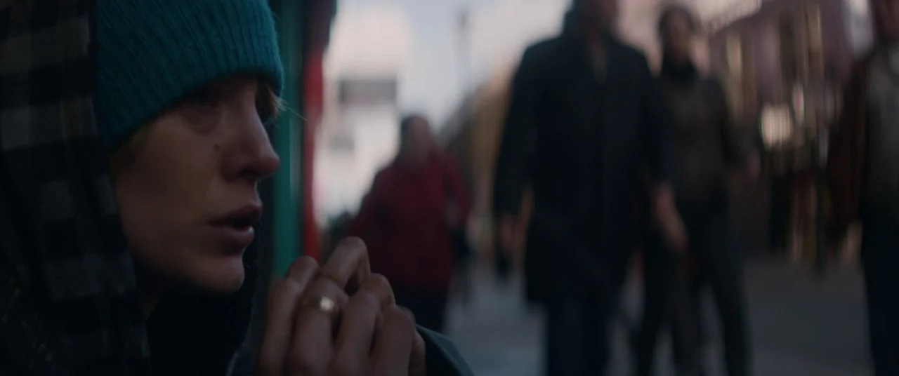 The Rhythm Section (2020) movie still: closeup — A blurred urban sidewalk shows pedestrians walking in the background behind a subject. A woman…