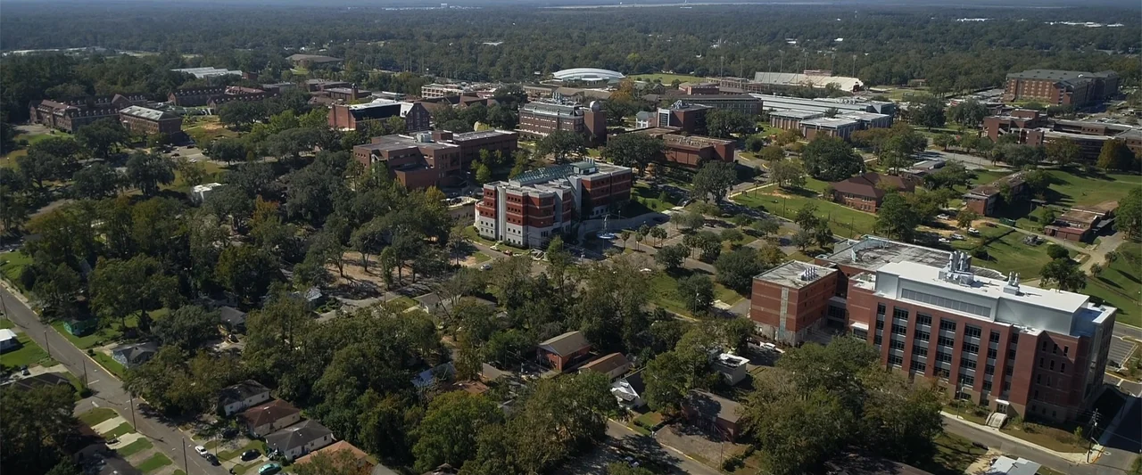 Init!ation (2021) movie still: establishing — An aerial view shows a sprawling university campus with brick buildings and dense trees. Wide streets…