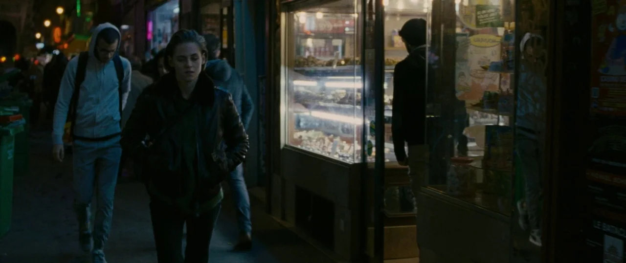 Personal Shopper (2016) movie still: medium — A nighttime sidewalk scene outside a small retail store with a lit display window. A young woman in a…