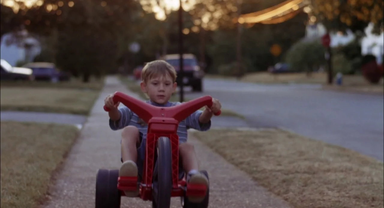 Trees Lounge (1996) movie still: medium — A residential sidewalk in late afternoon with parked cars blurred in the background. A young child rides a…