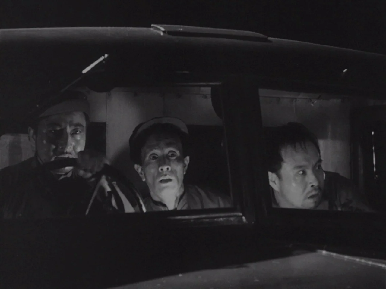 Godzilla Raids Again (1955) movie still: medium — The interior of a dark, cramped vehicle at night with three men sitting together. The men gaze…
