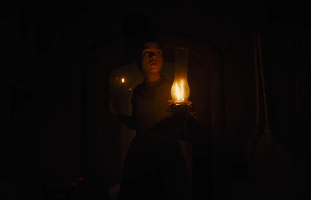 Gretel & Hansel (2020) movie still: medium — A dark, dimly lit corridor features an arched doorway and a wooden floor. A young girl holding a…