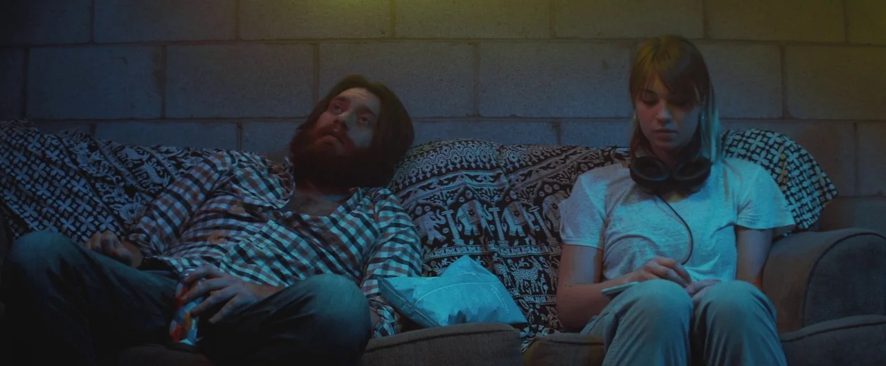 Poser (2021) movie still: medium — Two individuals sit on a sofa against a cinder block wall with patterned fabric. The man gazes upward while the…