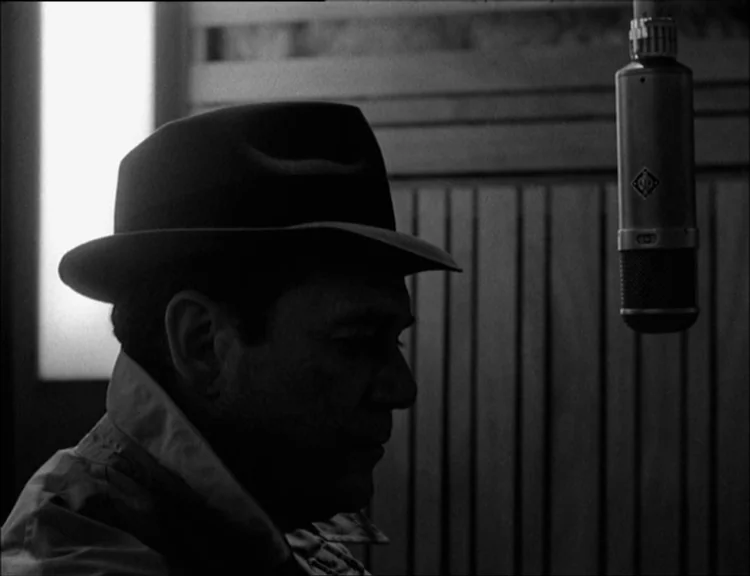 Alphaville (1965) movie still: closeup — A dark wooden-paneled room features a vintage studio microphone suspended from the ceiling. A man in a hat…
