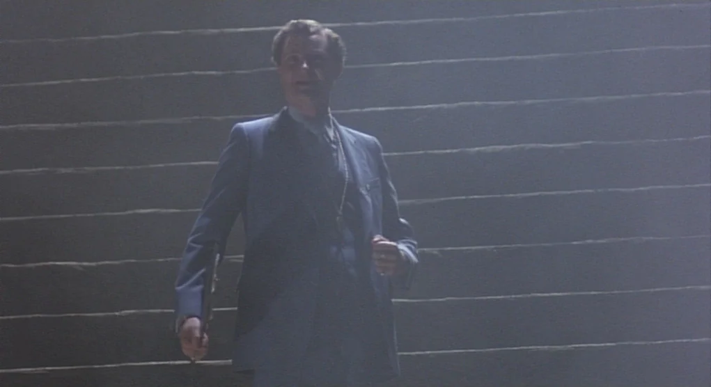 Time Bandits (1981) movie still: medium — A man stands on a wide stone staircase in a dimly lit, smoky interior. He wears a blue blazer and holds an…