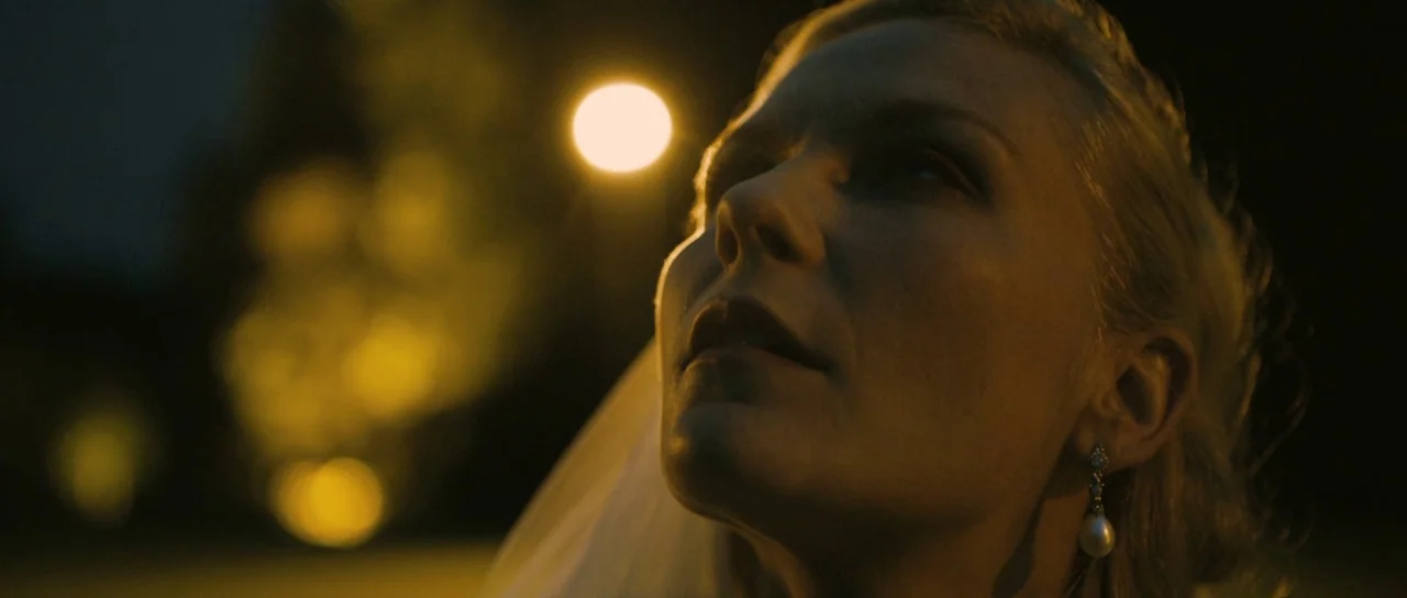 Melancholia (2011) movie still: closeup — A woman wearing a bridal veil looks upward at a bright, out-of-focus streetlamp. The dark background…