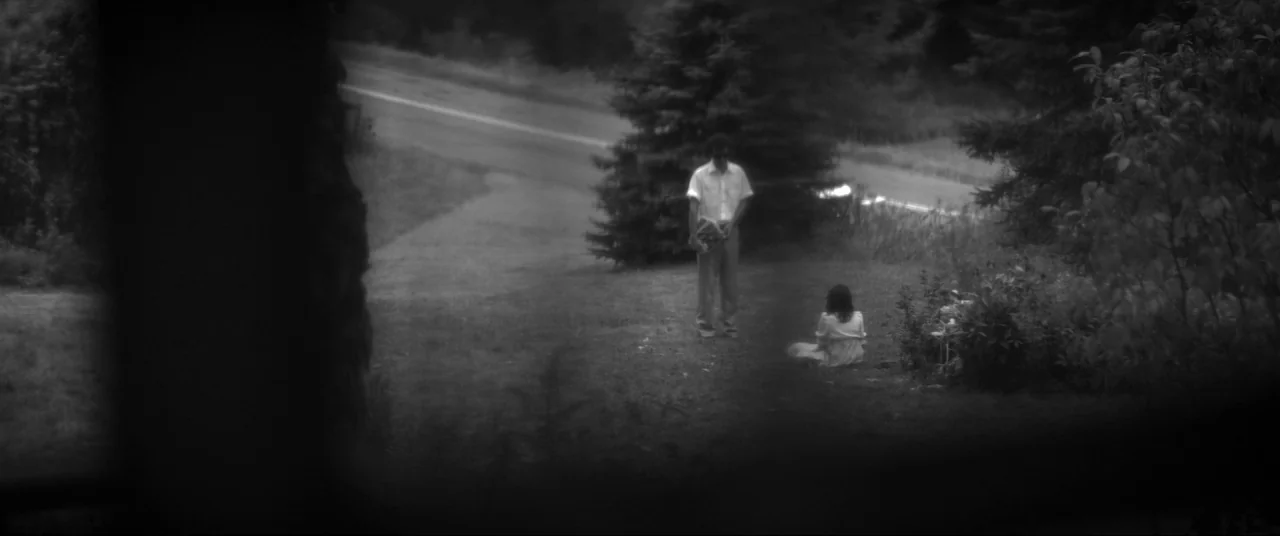 The Eyes of My Mother (2016) movie still: wide — A grassy yard bordered by dark evergreen trees sits beside a paved rural road. A man stands over a…