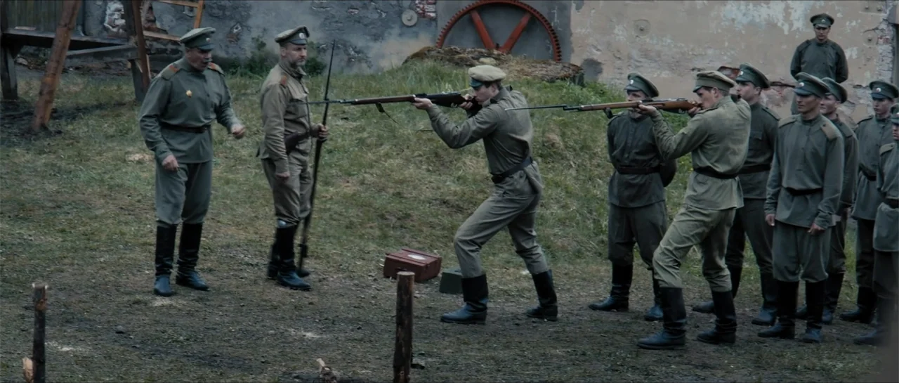 The Rifleman (2019) movie still: fullbody — An open dirt training ground features a stone wall and industrial machine parts behind. Several soldiers…