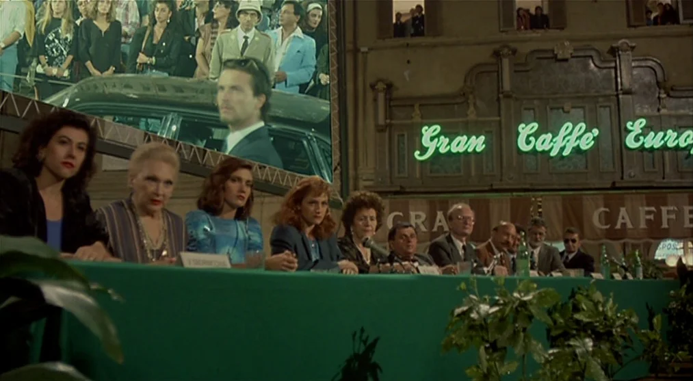 The Voice of the Moon (1990) movie still: wide — A group of people sits at a green table set up outside at night. A large screen displays a film…
