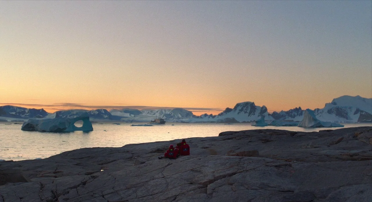 Where’d You Go Bernadette (2019) movie still: wide — A rocky coastal outcrop overlooks a calm bay filled with icebergs and distant mountains. Small…