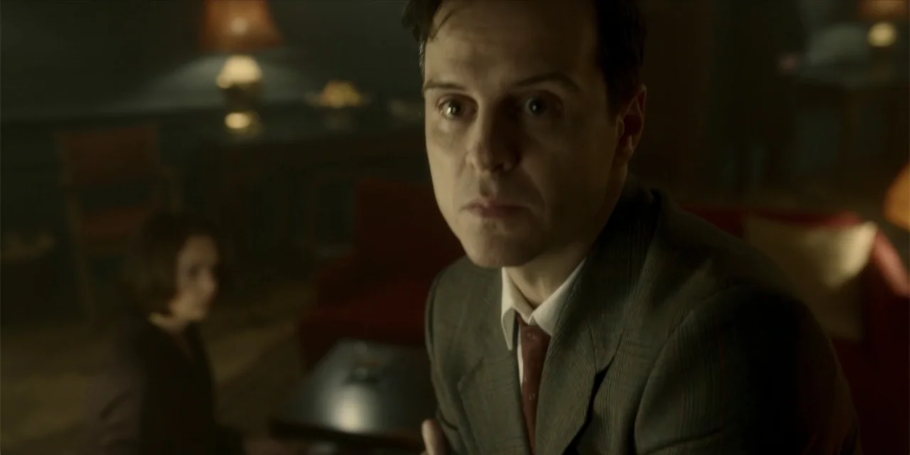 Oslo (2021) movie still: medium — A dimly lit room with vintage furniture and a dark red sofa. A man in a suit sits in the foreground while a woman…