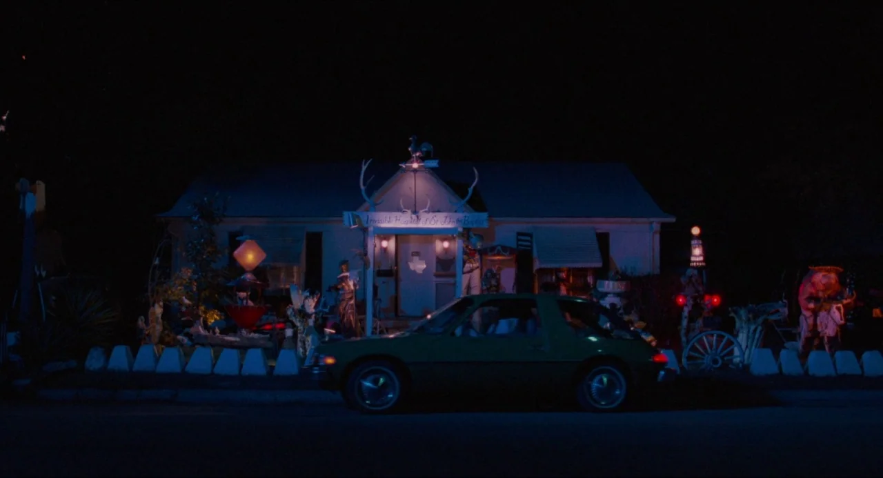 True Stories (1986) movie still: wide — A single-story residential structure is covered in dense, eclectic lawn art and ornaments. An older green…