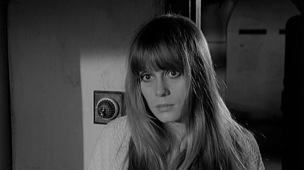 Cul-de-sac (1966) movie still: closeup — A dimly lit narrow space with a white wall and a vintage light switch. A young woman with long hair stands…