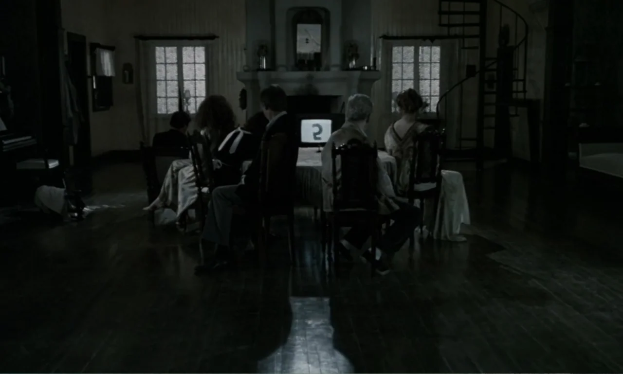 The Sacrifice (2015) movie still: wide — A dark, spacious room features a central television set surrounded by seated figures. Several people sit on…