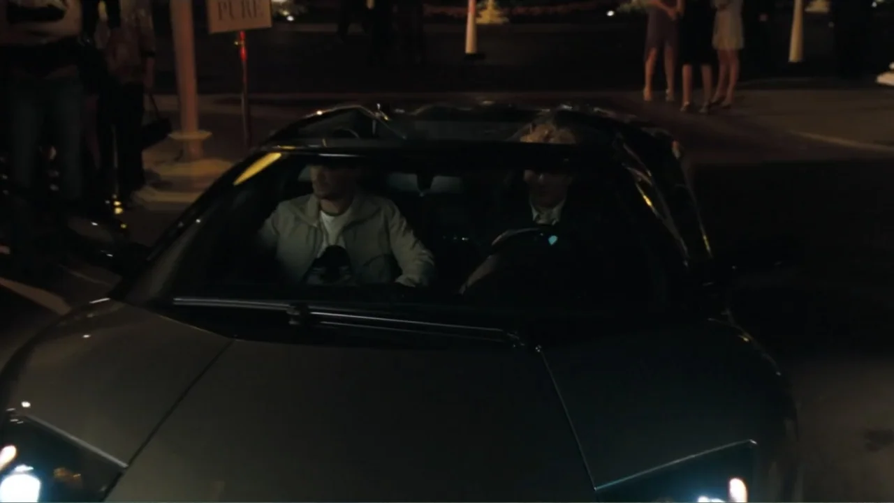 Redline (2007) movie still: medium — A dark convertible parked on asphalt at night with blurred figures in the distance. Two men sit inside the…