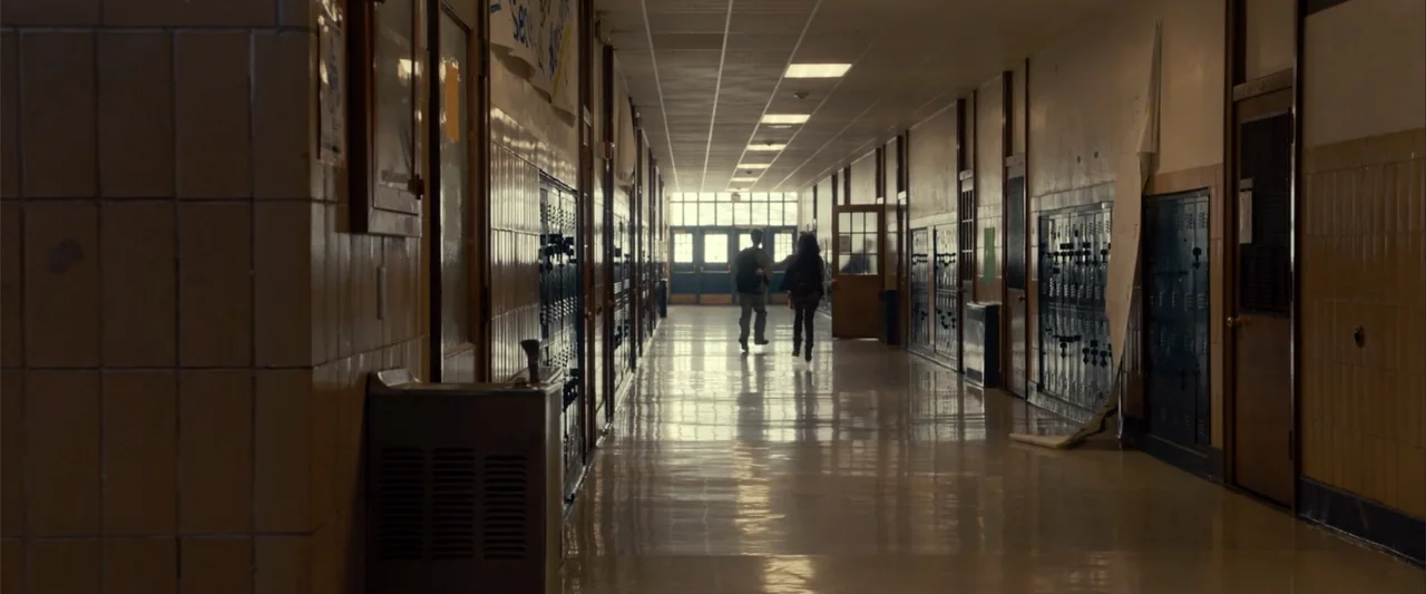 Frontera (2014) movie still: wide — A long institutional hallway with metal lockers lining the walls and polished tile floors. Two teenagers walk…
