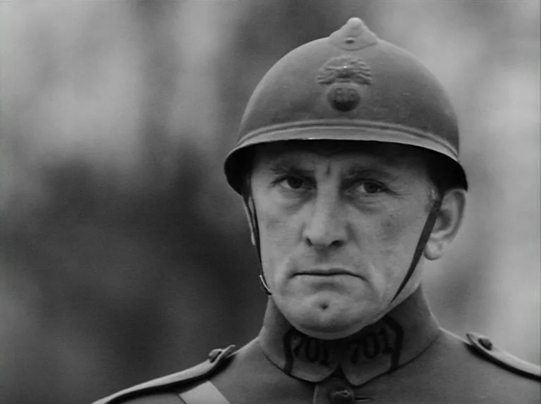 Paths of Glory (1957) movie still: closeup — A field with blurred outdoor background foliage typical of a military setting. A soldier in a helmet…