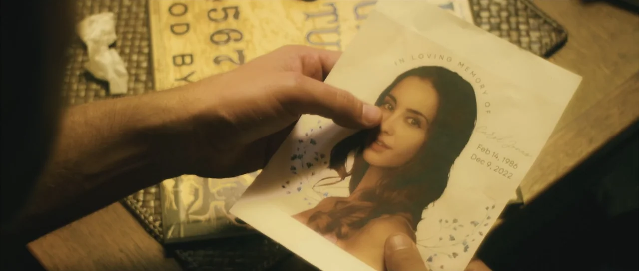 Night of the Missing (2023) movie still: closeup — A person holds a small memorial card with a woman's portrait and death dates. A portion of an…