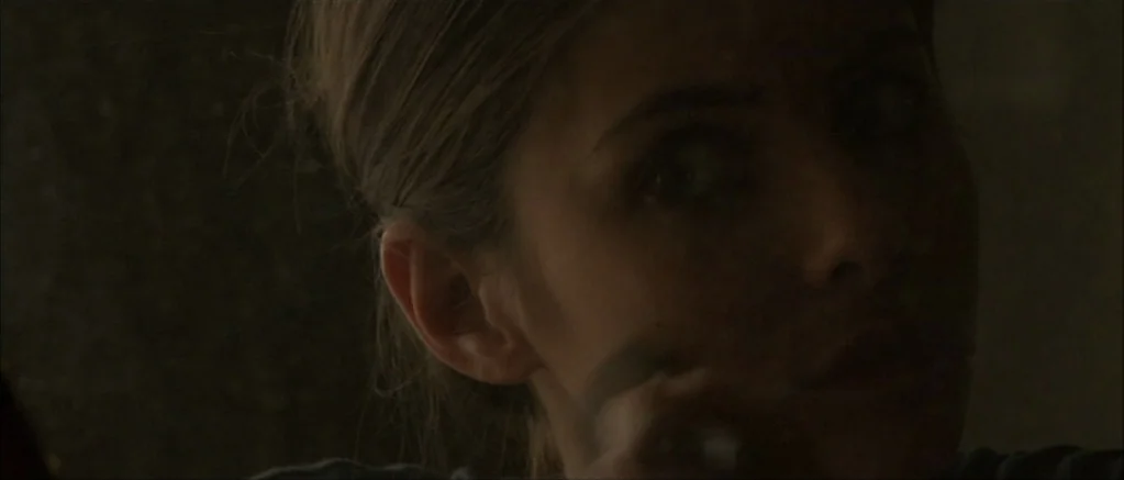 Genova (2008) movie still: closeup — A dimly lit interior space with shadows obscuring most of the surrounding area. A woman is positioned in the…
