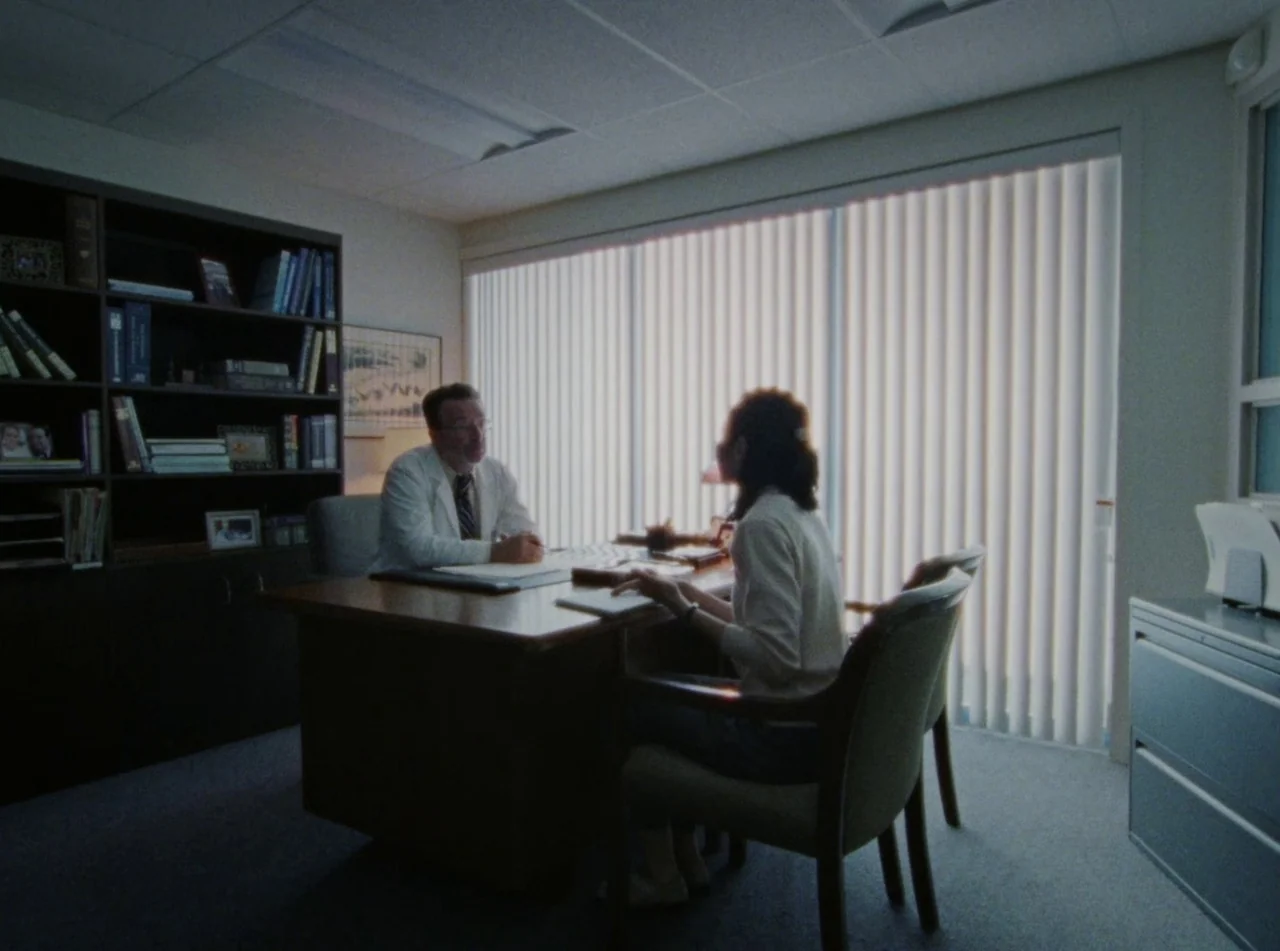 Riceboy Sleeps (2022) movie still: medium — A modest professional workspace with a bookshelf and a large wooden desk. A woman sits across from a man…