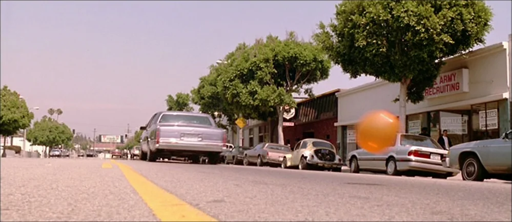Reservoir Dogs (1992) movie still: wide — An suburban street is lined with parked cars and storefronts under an overcast sky. An orange ball is…
