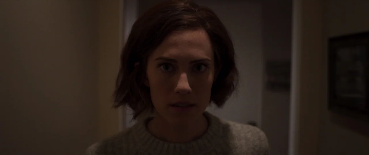 The Perfection (2018) movie still: closeup — A dimly lit indoor hallway with walls fading into deep shadow. A young woman faces forward looking…