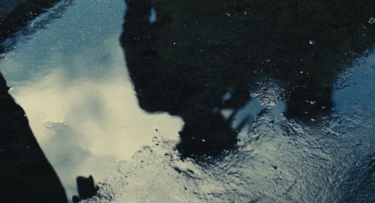 Past Lives (2023) movie still: closeup — A dark, textured asphalt ground contains a large puddle reflecting the overcast sky. Droplets hit the water…