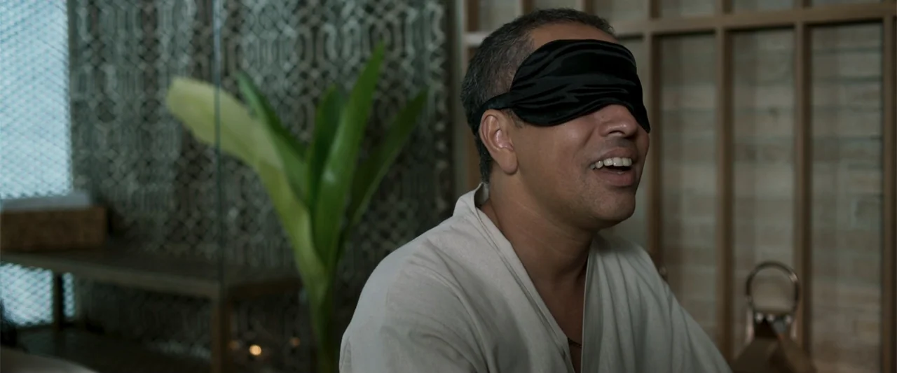 Férias Trocadas (2024) movie still: medium — A quiet spa interior features wooden lattice walls and a potted plant in background. A middle-aged man…