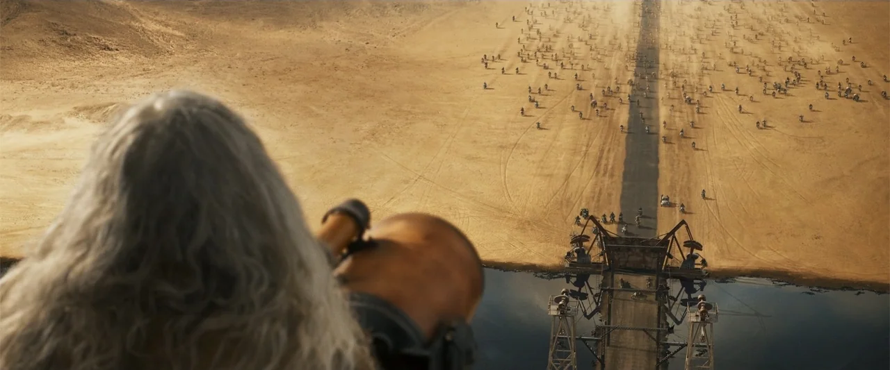 Furiosa: A Mad Max Saga (2024) movie still: wide — A vast, dry desert expanse features a long road leading toward a metal bridge structure. A large…