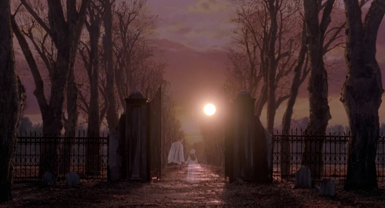 Hocus Pocus (1993) movie still: wide — A silhouette of two figures walking between stone pillars of a black iron gate. Tall leafless trees flank the…
