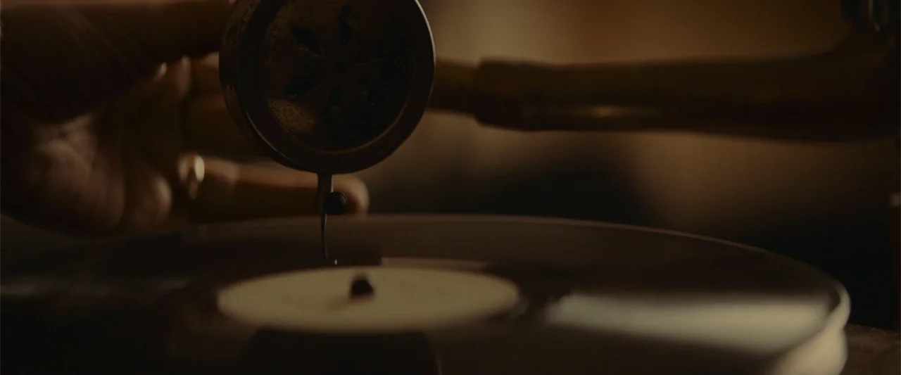 The Cellar (2022) movie still: closeup — A close view of a metal gramophone needle assembly above a dark vinyl record. The needle makes physical…