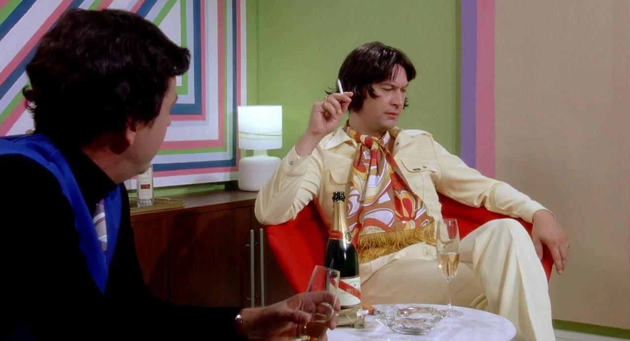 Viva (2007) movie still: medium — A 1970s living room features bold geometric wall art and colorful pastel decor. A man in yellow attire sits in a…