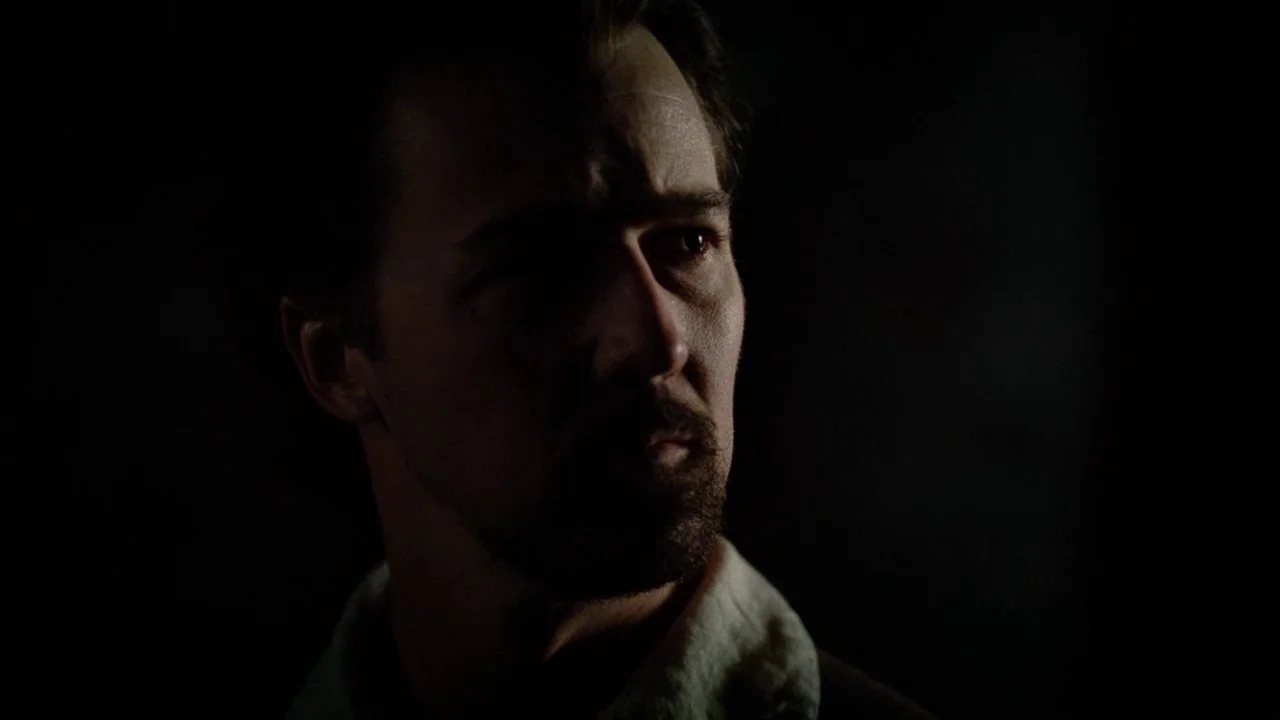 The Illusionist (2006) movie still: closeup — A man stands in a dark interior space with shadows obscuring the background. He is turned…