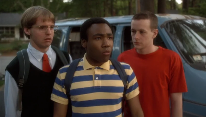 Mystery Team (2009) movie still: medium — A suburban residential setting with a blue van parked in the background. Three young men stand in a group,…