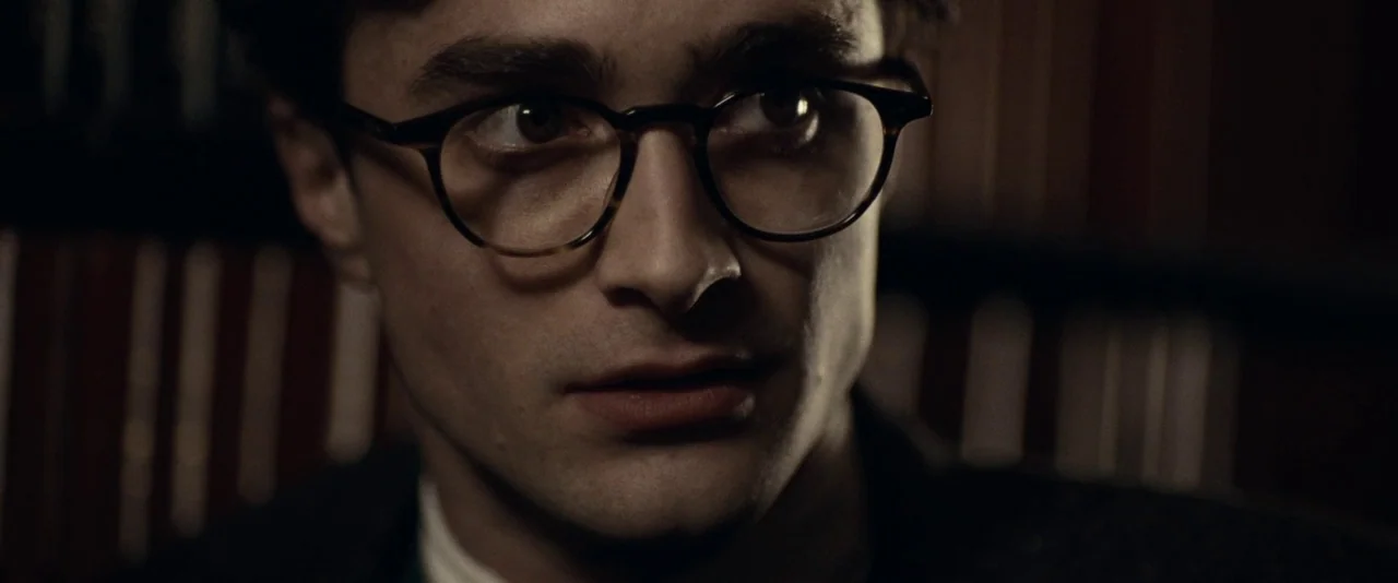 Kill Your Darlings (2013) movie still: closeup — A dimly lit interior space with shelves of books lining the wall in the background. A man wearing…