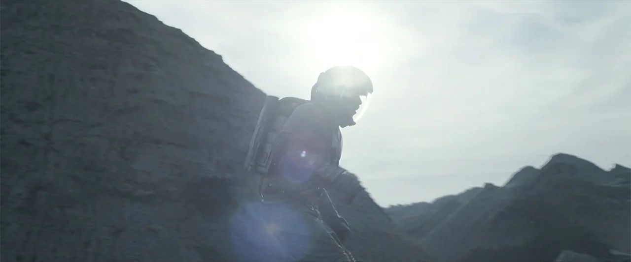 DISTANT (2026) movie still: medium — A figure in a protective space suit navigates a bleak, rocky, uneven terrain. The subject moves forward against…