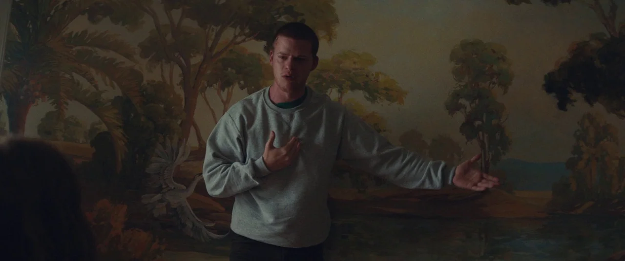 Honey Boy (2019) movie still: medium — A man stands in front of a wall featuring a large, painted mural of trees. He gestures with his hands while…