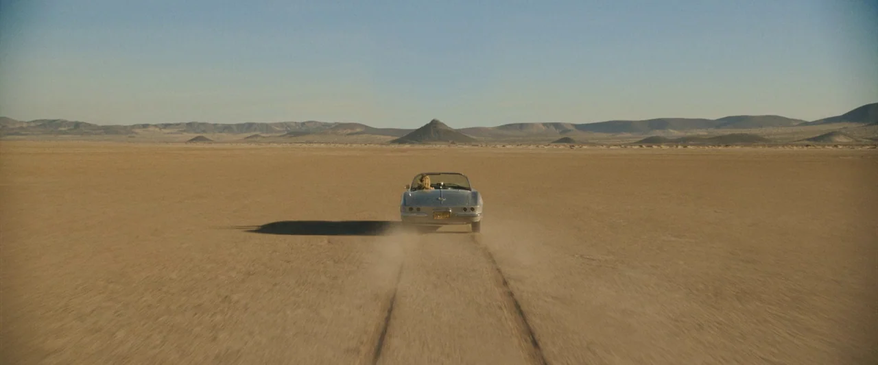 Don’t Worry Darling (2022) movie still: wide — A vast, flat, arid desert landscape features a single convertible driving into the distance. The…